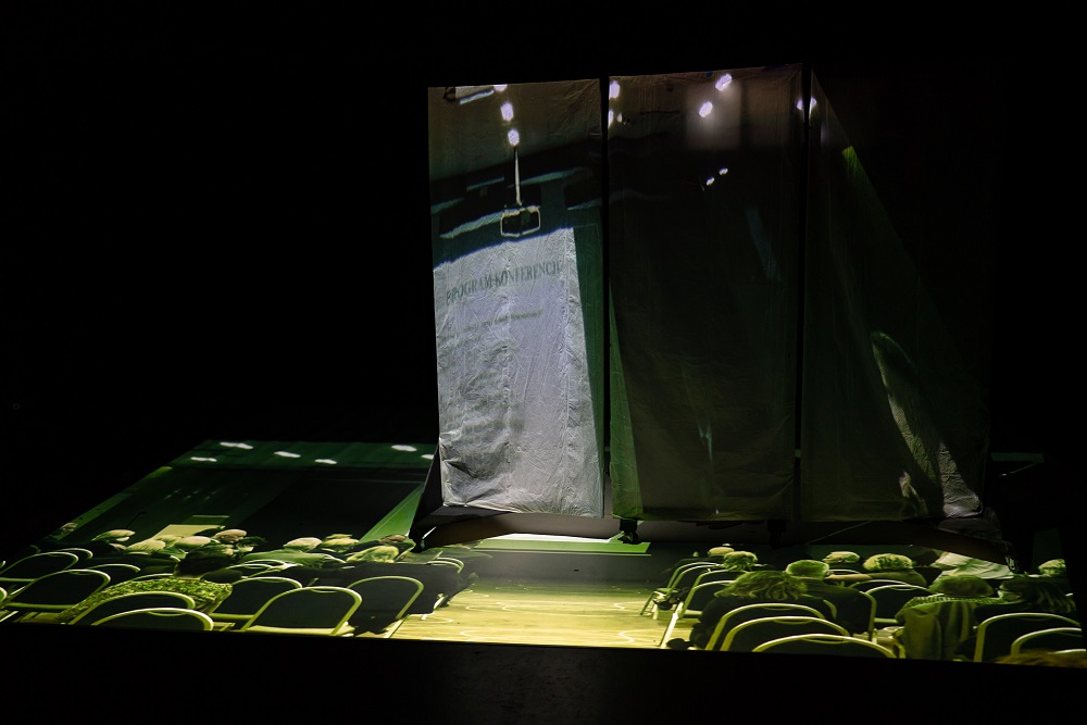 Rzut sceny z widownią oświetlonej na zielono, na scenie stroi parawan.
