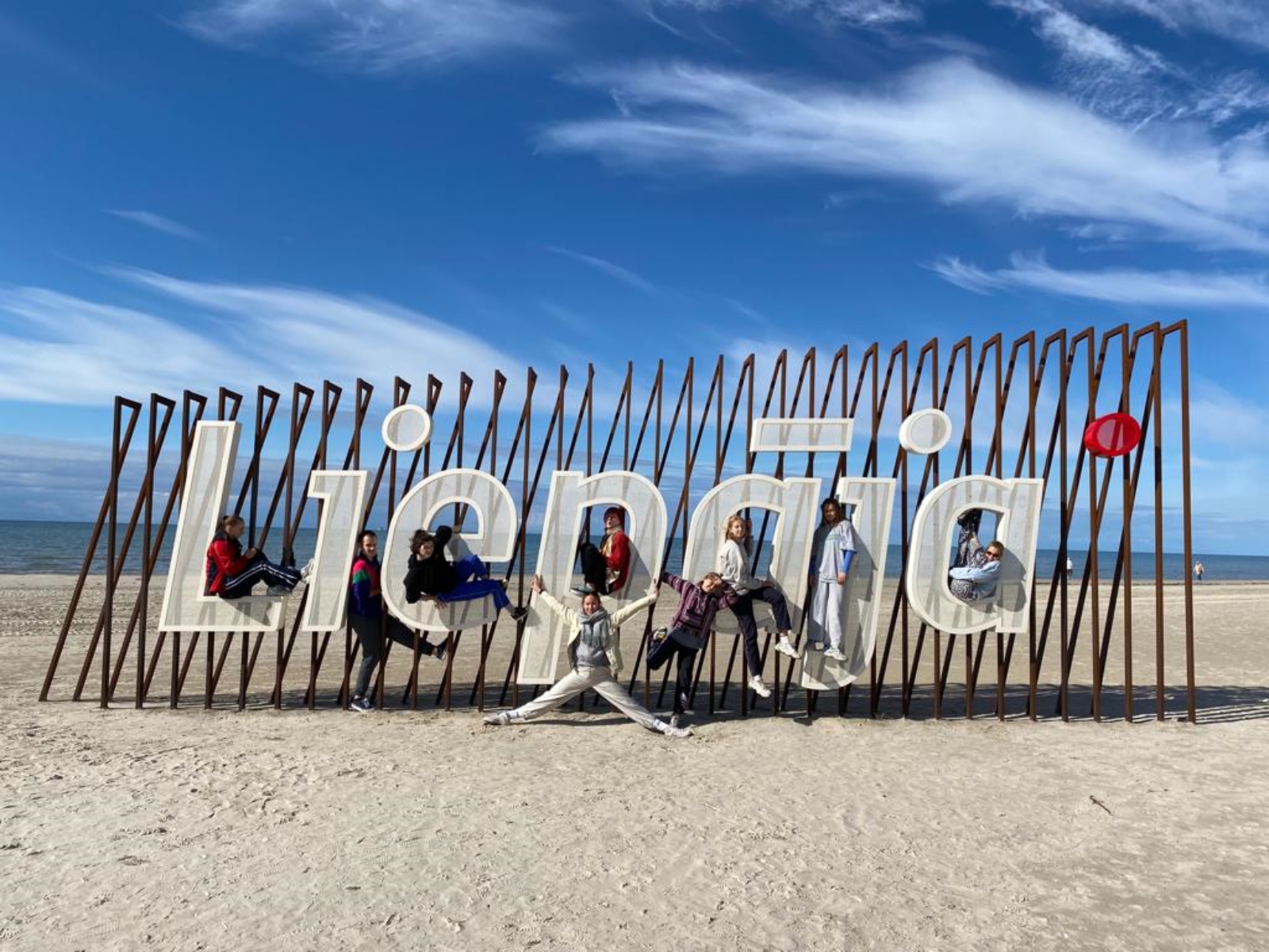 Grupa ludzi na plaży stojących przed tablicą z napisem Liepaja.