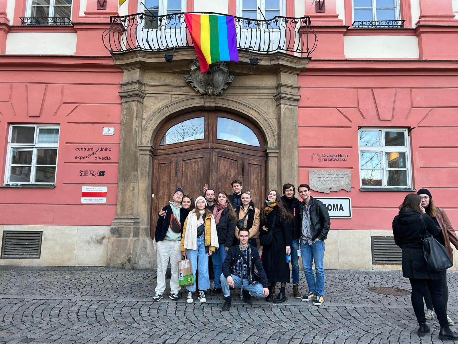 Grupa ludzi stoi przed budynkiem, na nimi na balkonie rozwieszona tęczowa flaga.