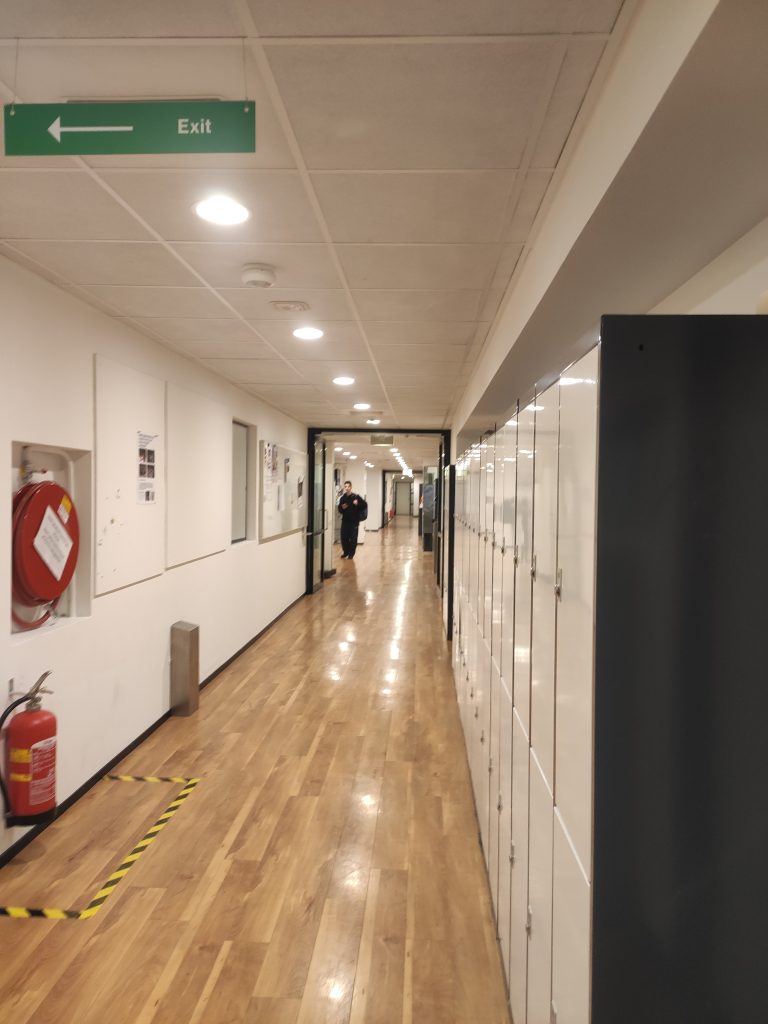 Długi rozświetlony korytarz o białych ścianach i drewnianej podłodze, po prawe stronie wzdłuż ścinany białe metalowe szafki studenckie, po lewej sprzęt przeciw pożarowy, na końcu korytarza postać ubrana na czarno.