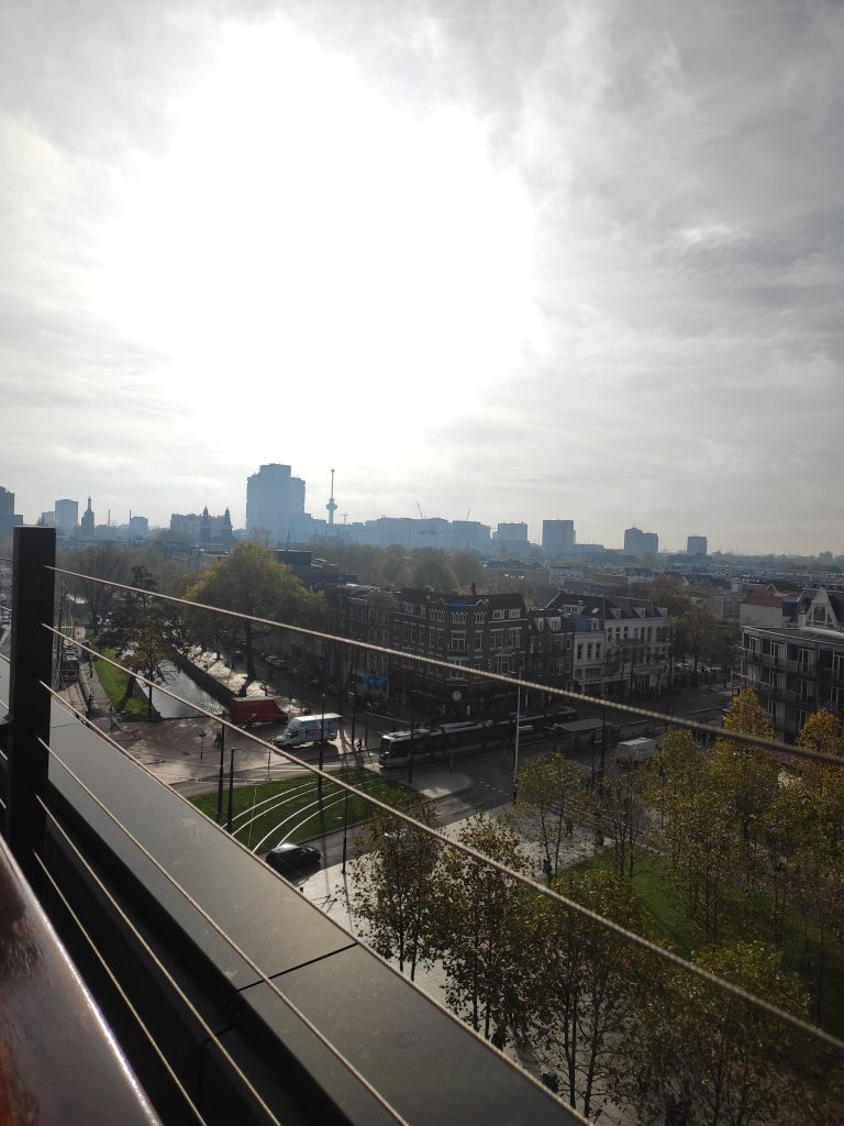 Panorama miasta widziana z balkonu, skrzyżowanie ulic, kamienice i kanał, w oddali ma horyzoncie wysokie nowoczesne budynki.