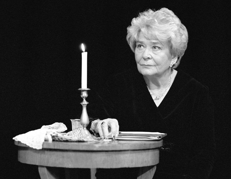 Danuta Michałowska na scenie, aktorka ubrana na czarno siedzi przy stoliku, na którym leżą drobne rekwizyty: rękawiczki, chusteczka, szklanka, na środku stoi srebrny świecznik a w nim paląca się świeca, w tle panuje półmrok.