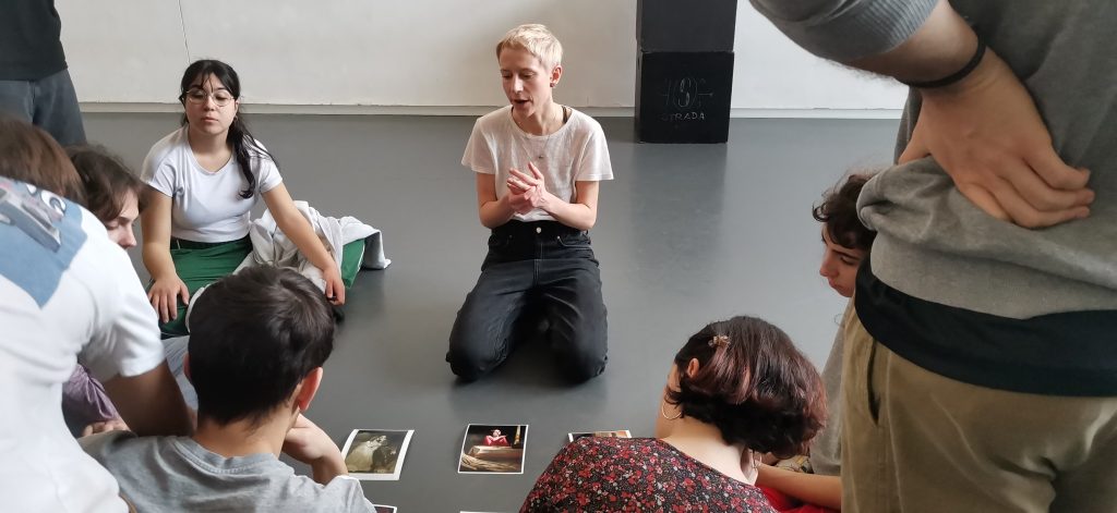 Grupa młodych ludzi siedząca wokół rozłożonych na szarej podłodze zdjęć, pośrodku młoda krótko ostrzyżona blondynka w jasnym T-shircie i czarnych dżinach.