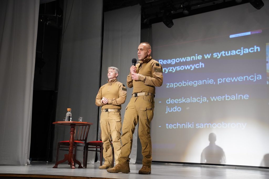 Dwóch funkcjonariuszy policji w mundurach stoi na scenie; jeden przemawia do mikrofonu, za nimi widać slajd z napisem „Reagowanie w sytuacjach kryzysowych”.