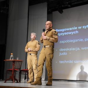 Dwóch funkcjonariuszy policji w mundurach stoi na scenie; jeden przemawia do mikrofonu, za nimi widać slajd z napisem „Reagowanie w sytuacjach kryzysowych”.