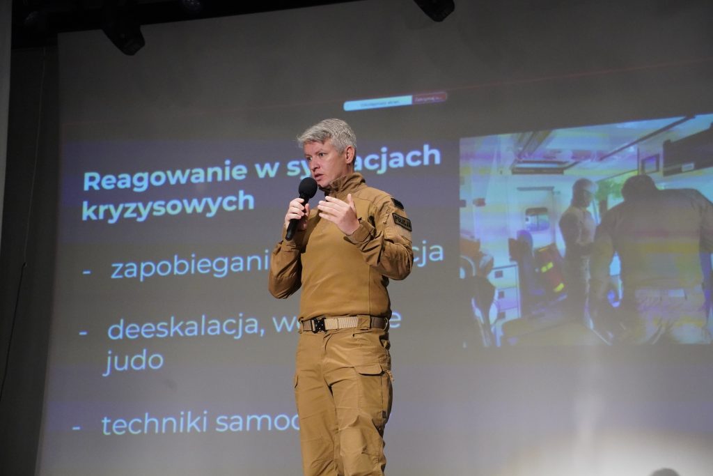 Policjant w brązowym mundurze przemawia do mikrofonu na tle slajdu prezentacyjnego przedstawiającego działania interwencyjne.