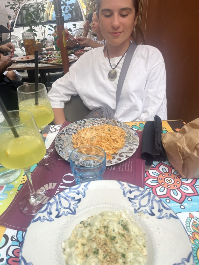 Osoba siedząca przy restauracyjnym stoliku z talerzami makaronu i napojami.