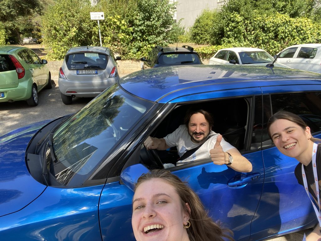 Uśmiechnięta grupa robiąca wspólne zdjęcie przy niebieskim samochodzie na parkingu.