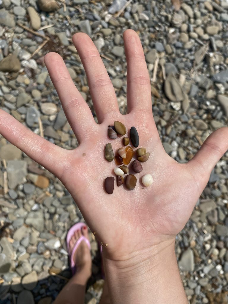 Otwarta dłoń trzymająca kilkanaście drobnych, kolorowych kamyków na tle kamienistej plaży.