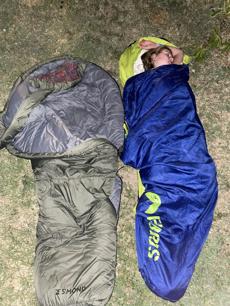 Osoba śpiąca nocą pod gołym niebem w niebieskim śpiworze; obok leży rozłożony szarozielony śpiwór.