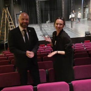 Prezydent Wrocławia Jacek Sutryk oraz prorektorka AST Ewelina Ciszewska podczas rozmowy na tle teatralnej sceny. Atmosfera luźna i życzliwa, oboje uśmiechnięci, za nimi przestrzeń sceniczna w trakcie przygotowań.