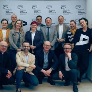 Zdjęcie grupowe kilkunastu osób stojących i siedzących przed ścianką z napisem „Akademia Sztuk Teatralnych”. Uczestnicy pozują do zdjęcia, ubrani głównie w stroje półformalne i formalne.