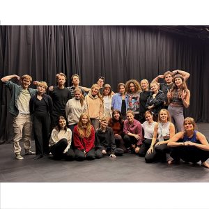 Grupa około dwudziestu młodych osób pozuje razem na scenie w sali teatralnej, na tle czarnej kurtyny. Część stoi, część siedzi na podłodze; niektórzy robią żartobliwe gesty, jak napinanie mięśni czy znak pokoju. Wszyscy są ubrani swobodnie i patrzą w stronę aparatu.