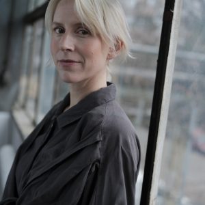 „Portret dr Sary Celler-Jezierskiej stojącej przy dużym oknie. Ubrana w ciemnoszarą koszulę, patrzy w obiektyw z delikatnym uśmiechem. W tle widoczne są rozmyte elementy miejskiej zabudowy i wnętrze z przeszkloną ścianą