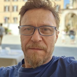 „Portret dr. hab. Grzegorza Wojdona w okularach, ubranego w granatową koszulkę. Zdjęcie wykonane na tle zabytkowej zabudowy miejskiej z rozmytym rynkiem w tle.”