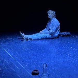 2. Performer siedzi na pustej, ciemnej scenie oświetlonej niebieskim światłem; przed nim stoją szklana karafka i popielniczka.