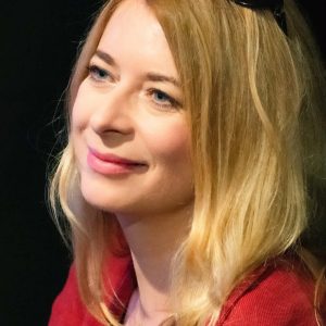 Ewa Kaim, uśmiechnięta blondynka w czerwonym żakiecie, sfotografowanej z profilu na ciemnym tle. Na głowie ma okulary przeciwsłoneczne.